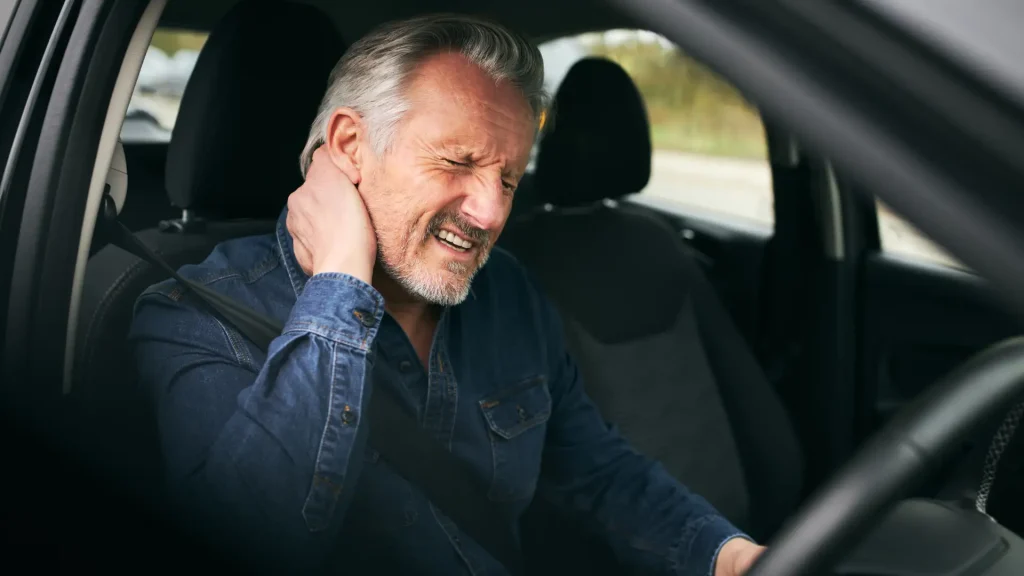 An elderly man holding his neck in pain while sitting in a car, possibly after a car accident.