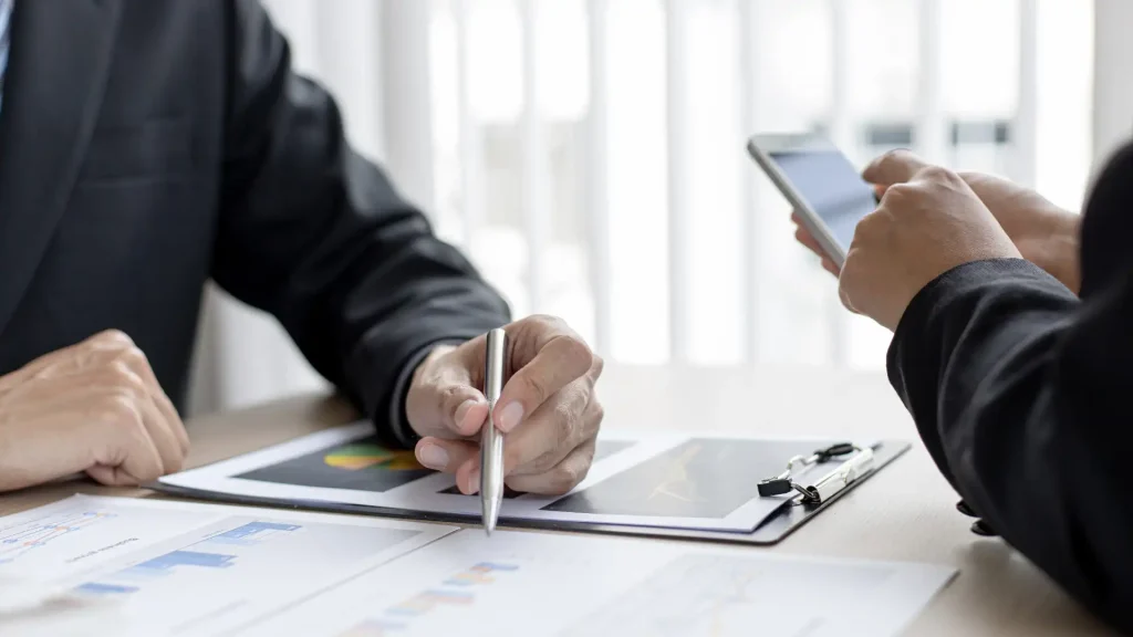 A person holding a pen, reviewing documents with graphs, while another person uses a smartphone.