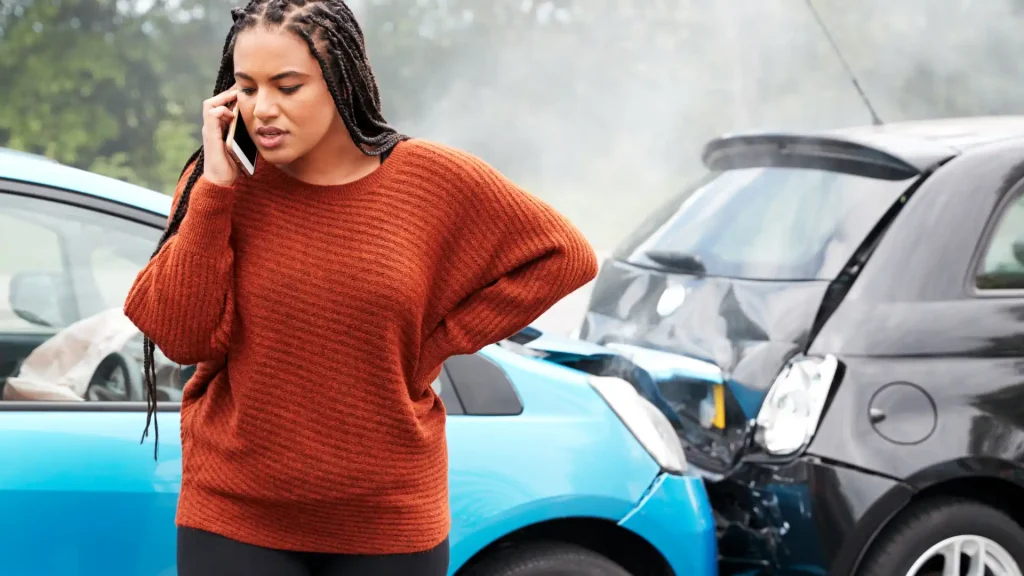 A woman in a red sweater standing next to a car accident, holding a phone, likely calling for help after the crash